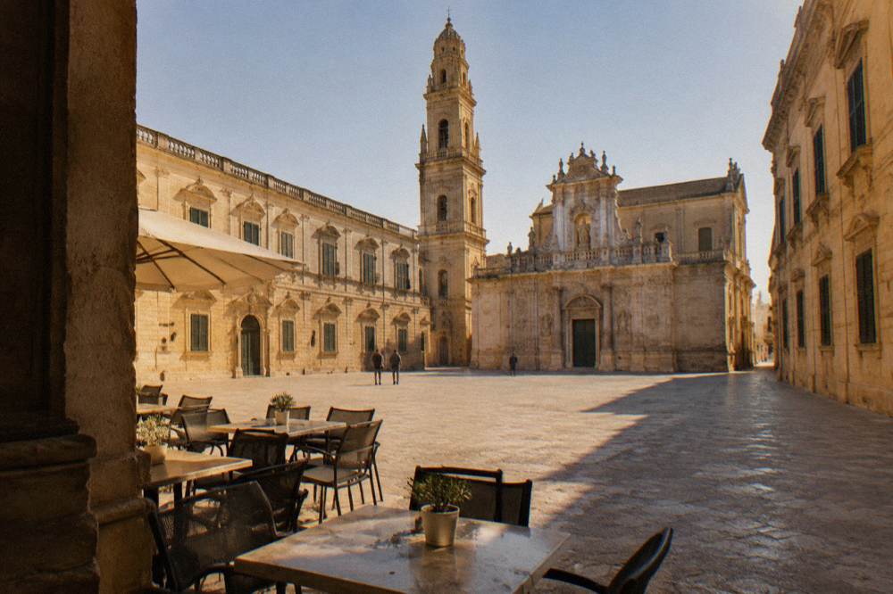 Empty café tables on a sunlit stone square in the early afternoon, with shuttered windows behind them.