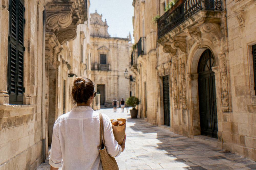 A person carrying a paper bag of pastries along a narrow limestone street between tall baroque buildings.