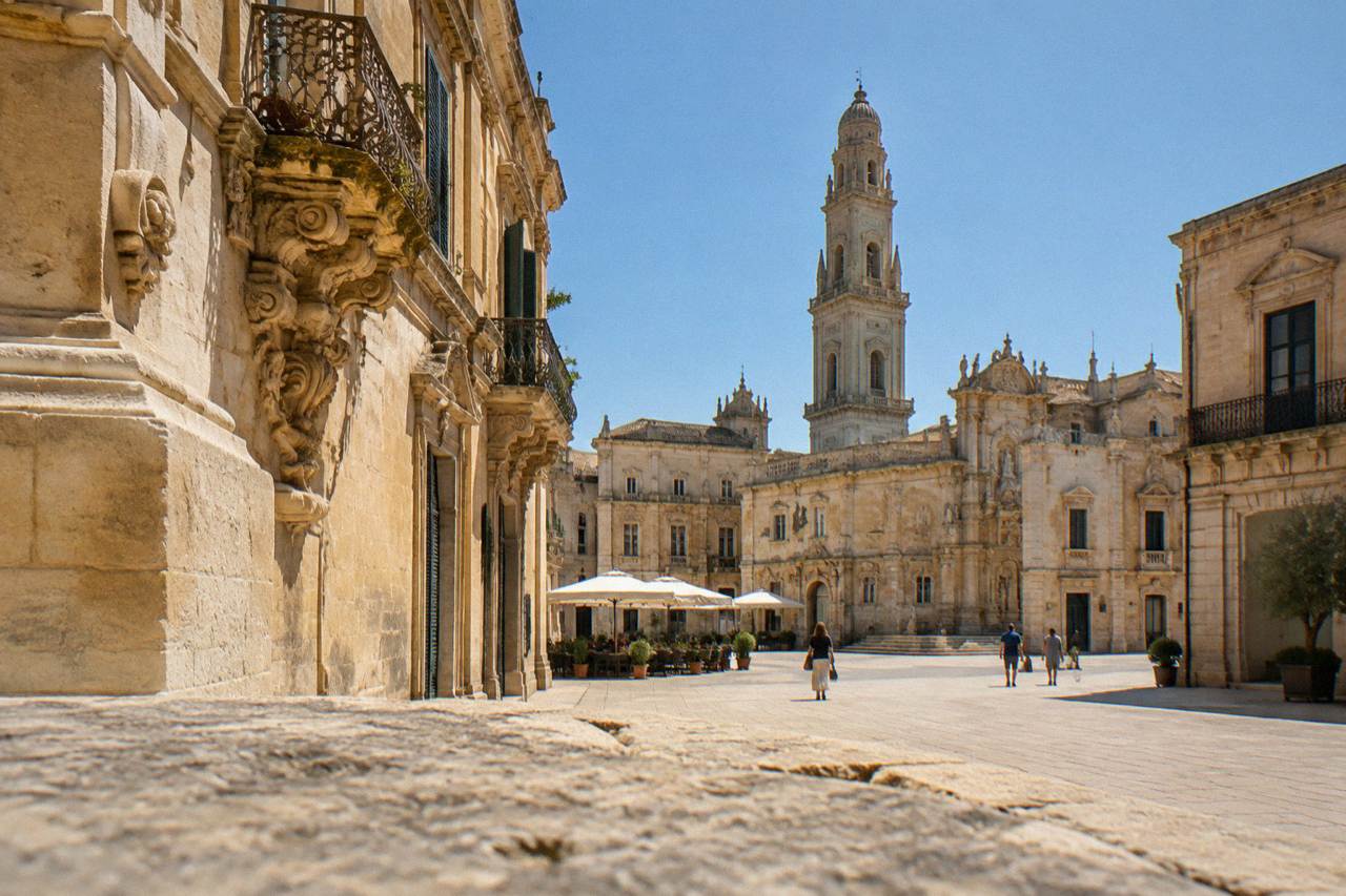 A sunlit Lecce square with honey-coloured baroque facades and a tall bell tower under a clear sky