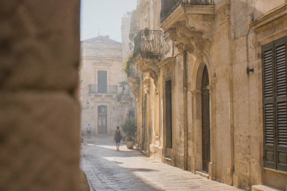 A narrow Lecce street in golden late afternoon light with stone balconies and closed shutters