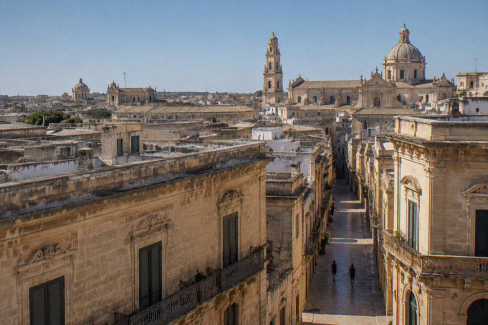 A wide view of Lecce rooftops and church domes under a clear blue sky at midday