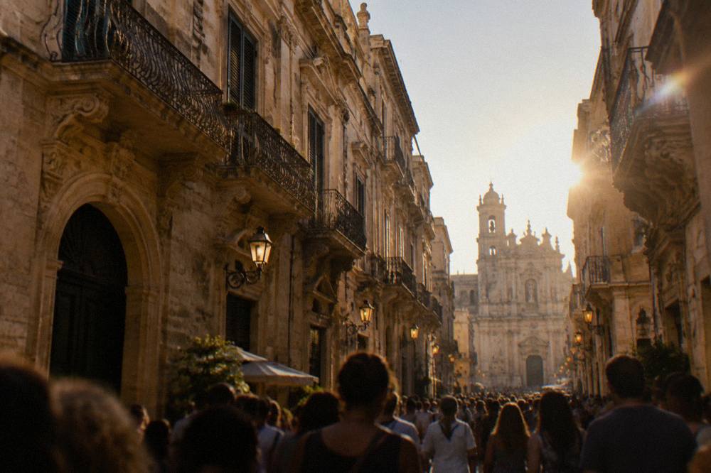 Dense evening crowds filling a lamp-lit baroque street
