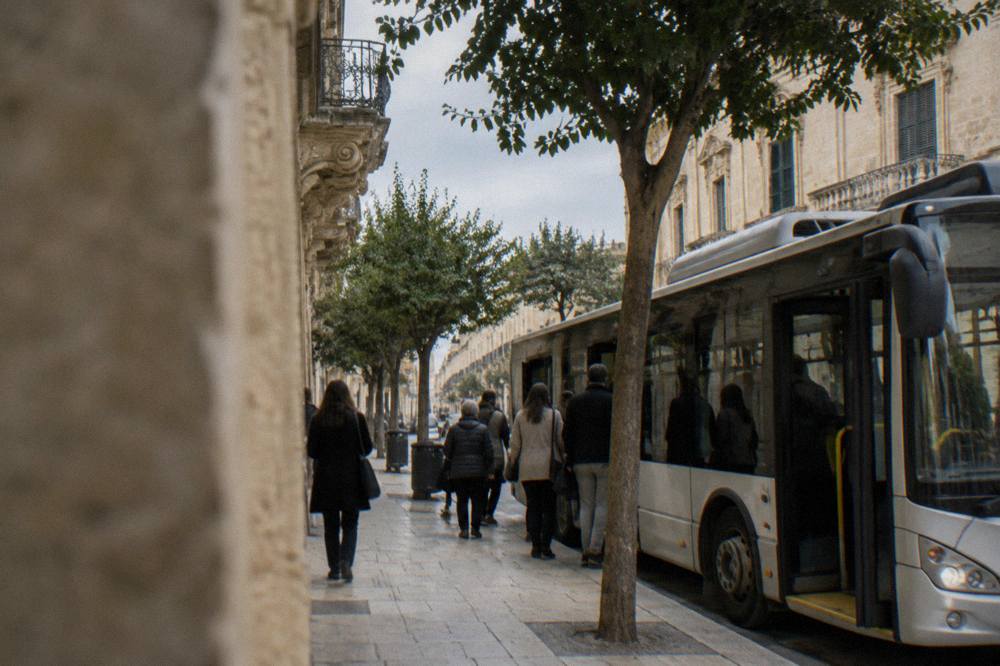 A city bus parked on a tree-lined street in Lecce with passengers boarding