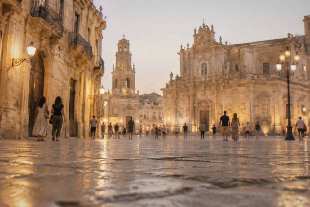 A floodlit Lecce square at dusk with people strolling under warm lamps beside baroque buildings