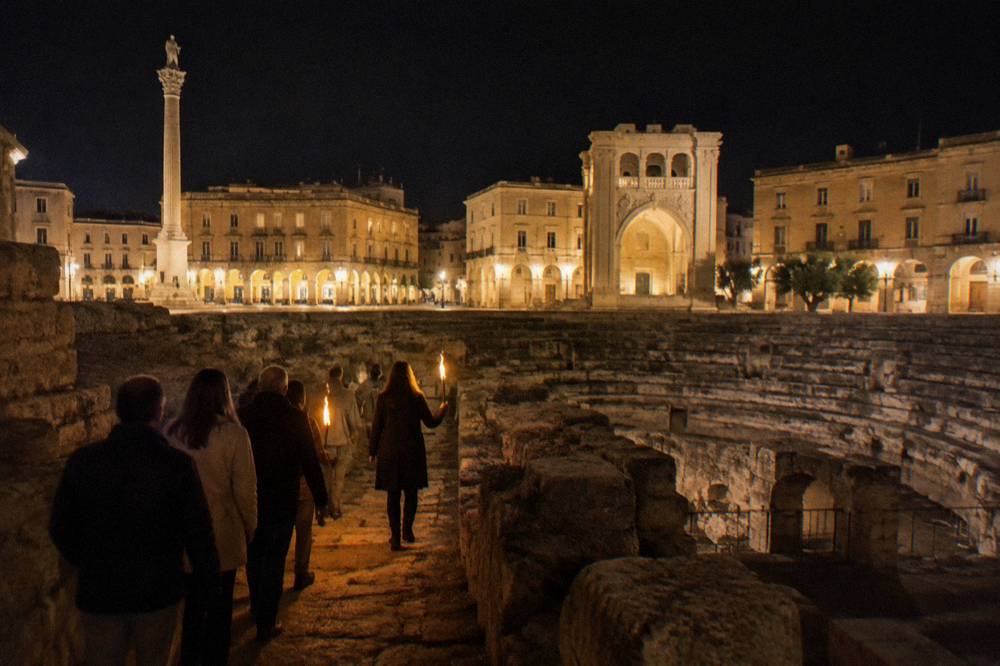 A guide leading a small group with torches through ancient stone ruins at night