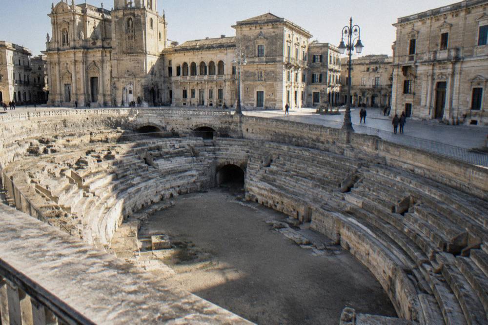 Roman amphitheatre remains in a paved square beside baroque buildings
