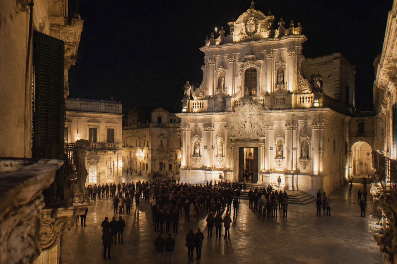 A floodlit baroque church facade in Lecce at night with crowds gathered in the square below