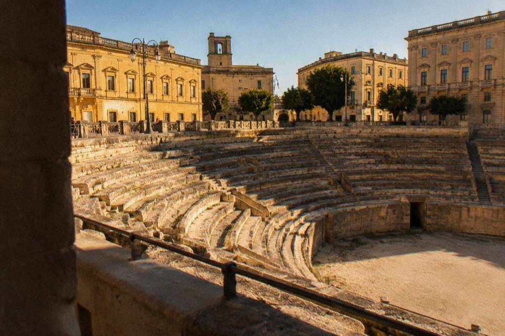 An elliptical sunken Roman amphitheatre framed by golden buildings on a city square