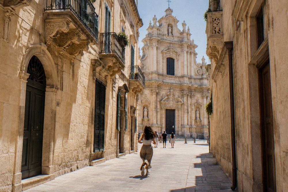 A towering baroque church facade of pale golden stone seen from a narrow street