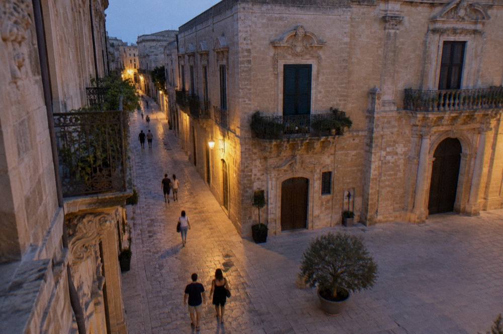 People walking on pale stone paving past baroque buildings in warm evening light