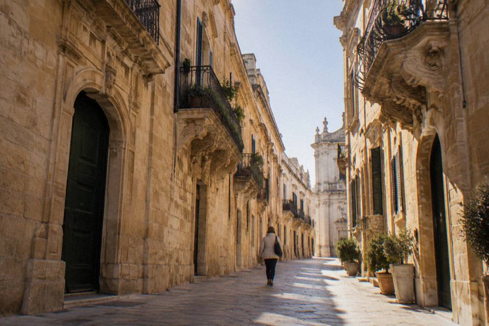 A person in a light jacket walking a narrow baroque street in Lecce in autumn