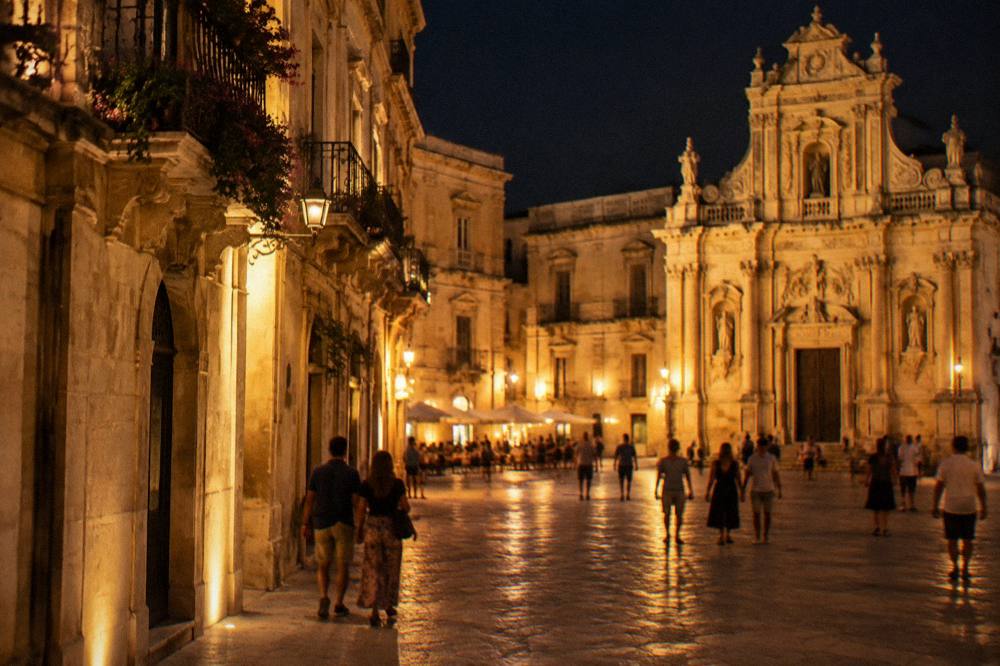 A lit-up baroque square in an Italian town at night with people strolling between historic buildings