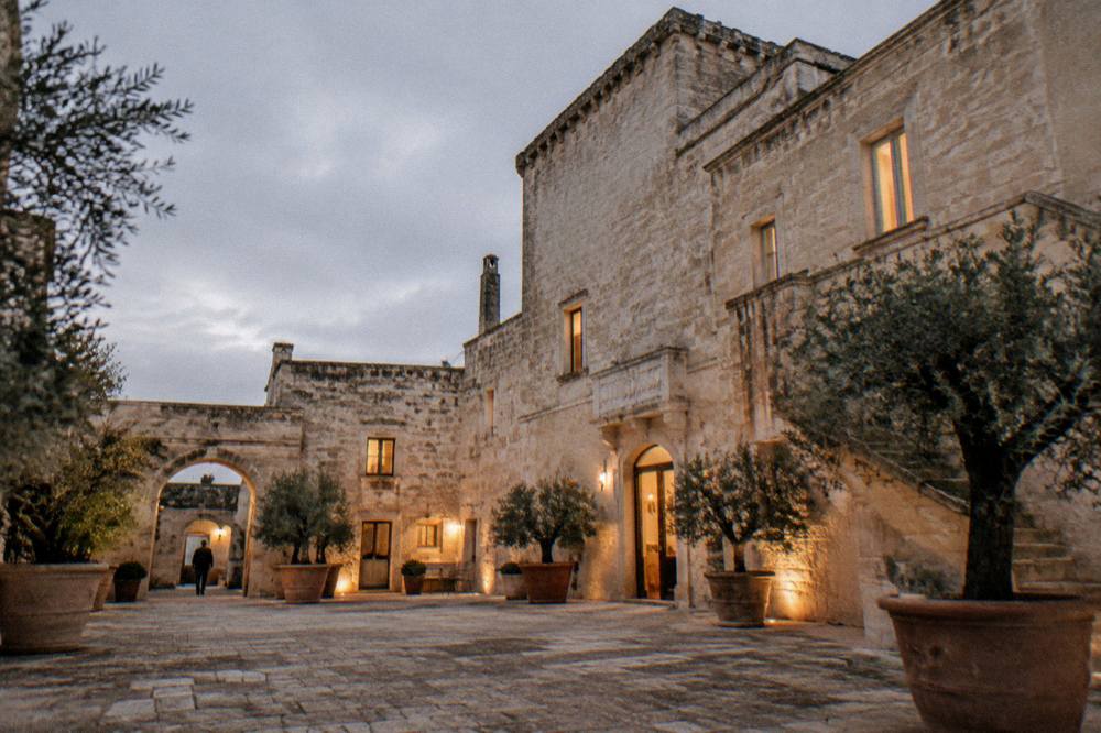 A stone masseria courtyard at dusk with lit windows and potted olive trees
