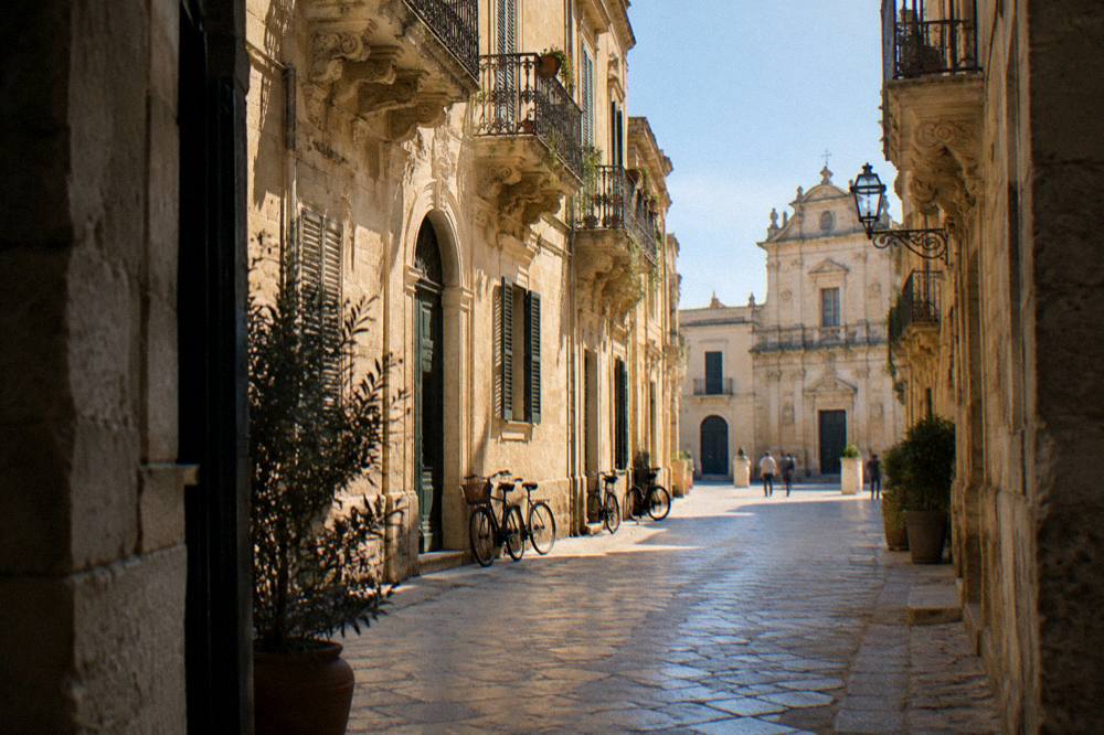 A sunlit Italian old town street with baroque stone facades and parked bicycles