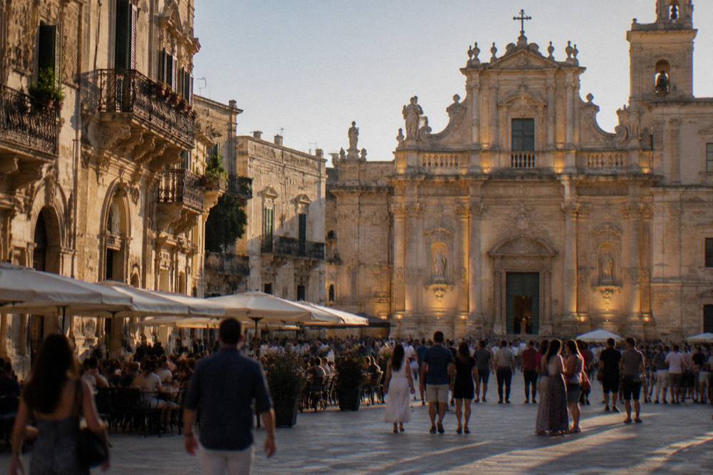 A crowded summer evening square in an Italian baroque city with illuminated church facades