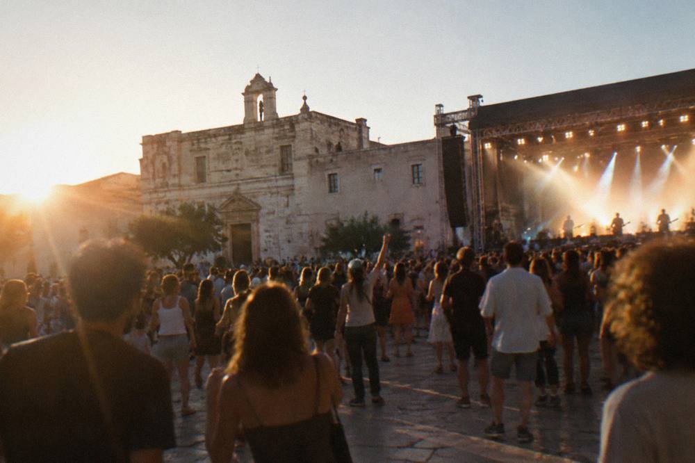 A night crowd dancing in front of a brightly lit festival stage in a southern Italian town