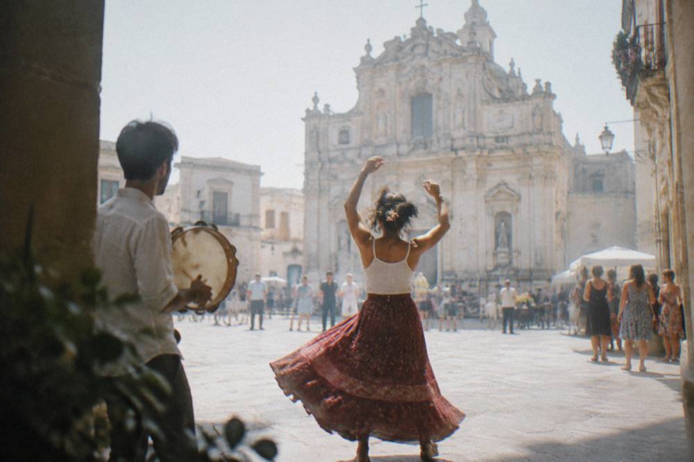 A dancer spinning with arms raised while a tambourine player keeps rhythm at a village feast