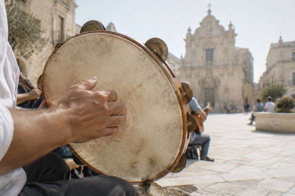 A close view of hands playing a frame drum with cymbals in its rim at a folk music session