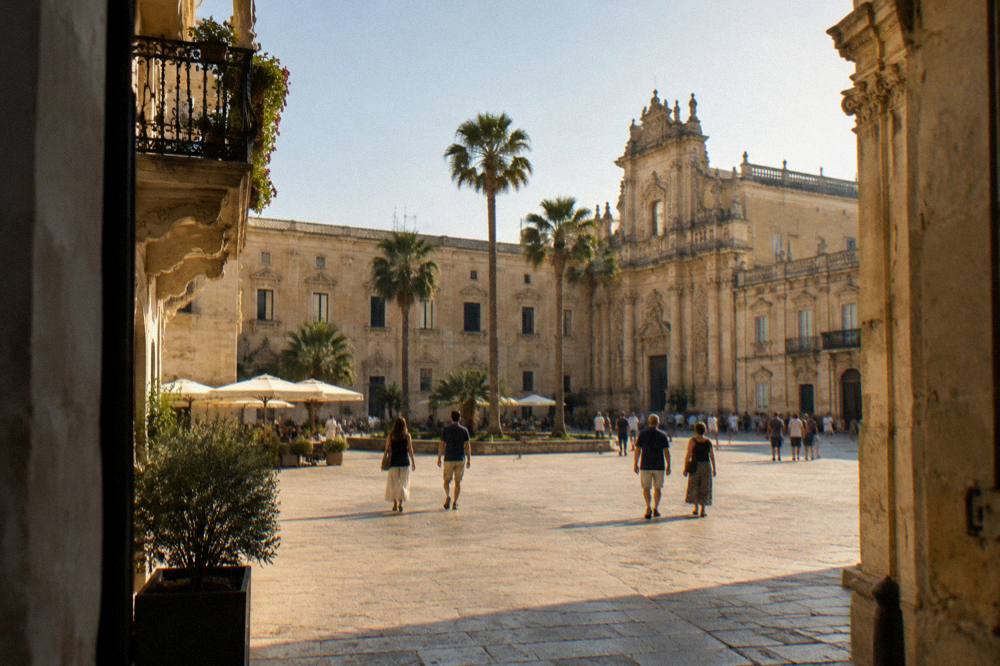 A wide square in Lecce with baroque buildings, palm trees and people walking in warm evening light