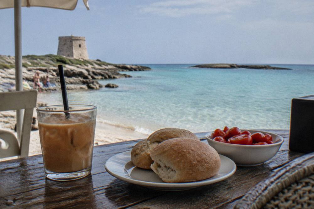 A shaded table with iced coffee, bread rolls and tomatoes at a beach bar