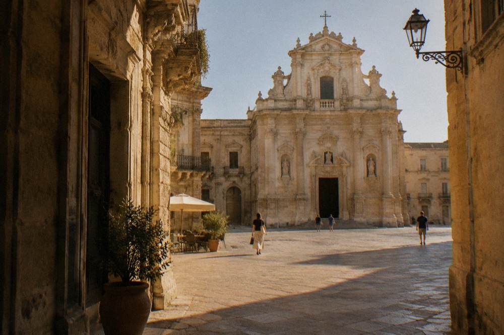 A baroque church facade of pale golden stone seen from a sunlit square