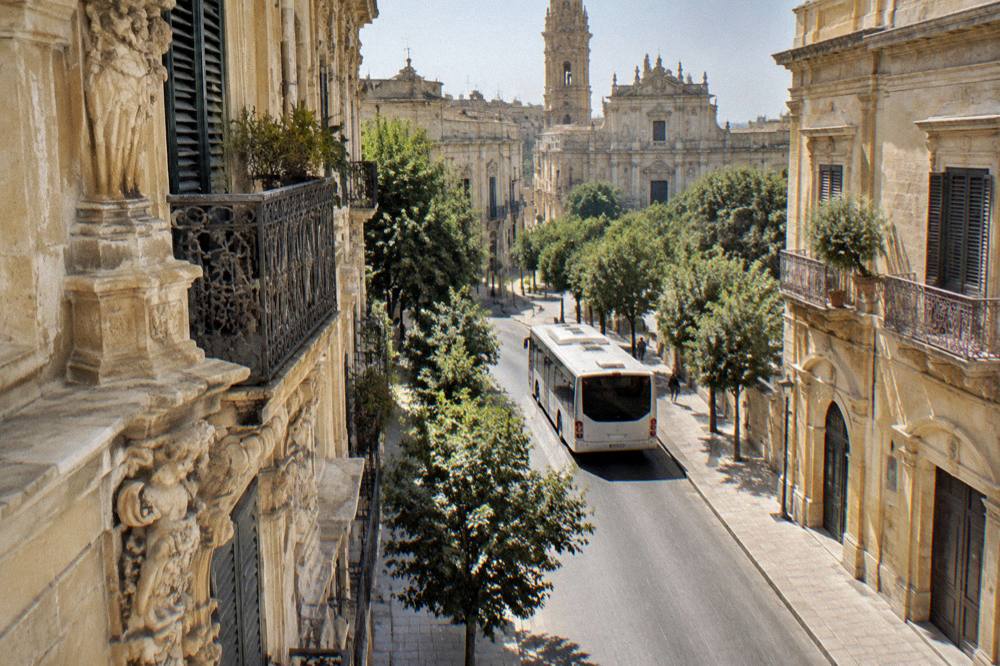 A city bus pulling away from a stop on a tree-lined street in a southern Italian town