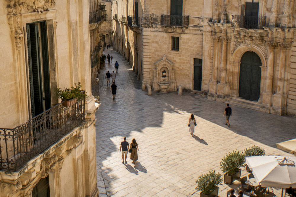 A stone square in an old Italian town with people walking between baroque buildings in late afternoon light