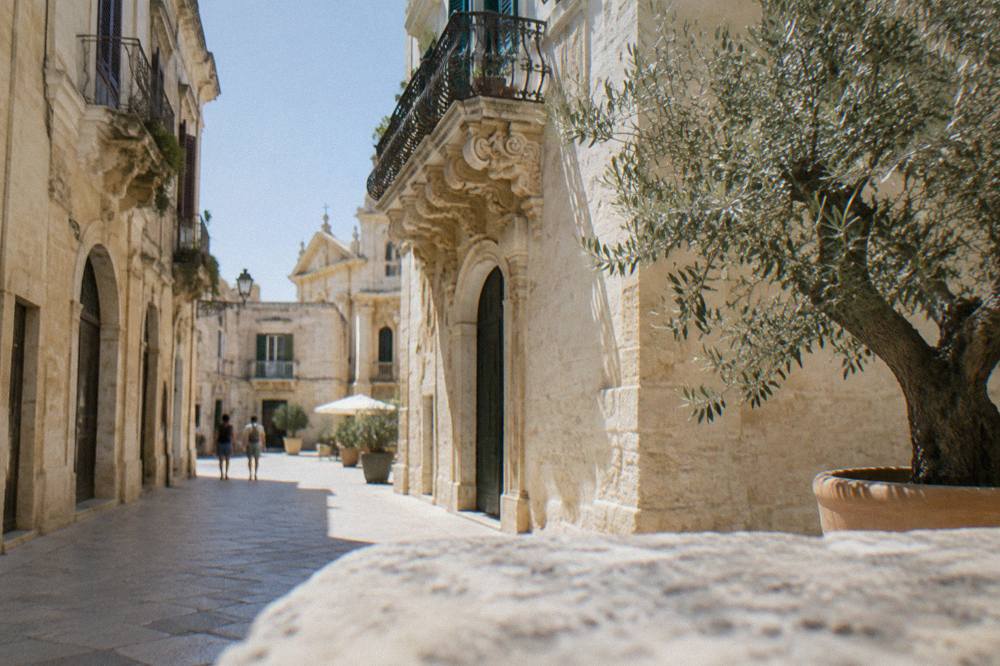 A white-washed Salento town street with a baroque balcony and olive tree