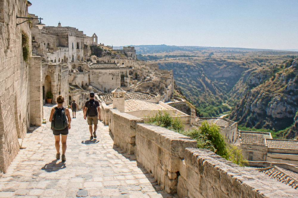 Travellers with backpacks walking a stone street toward the open countryside beyond the old town