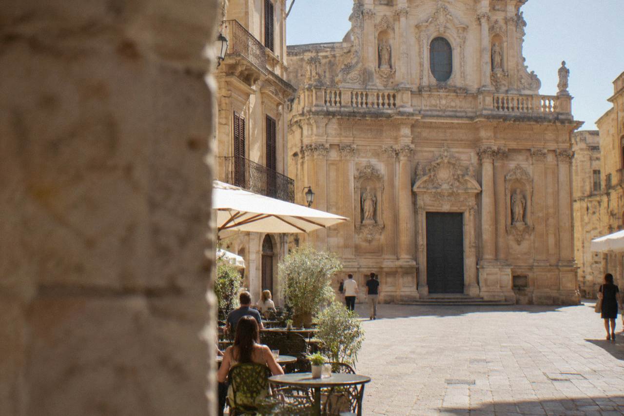 A golden limestone square in Lecce at morning with a baroque church facade rising above cafe tables
