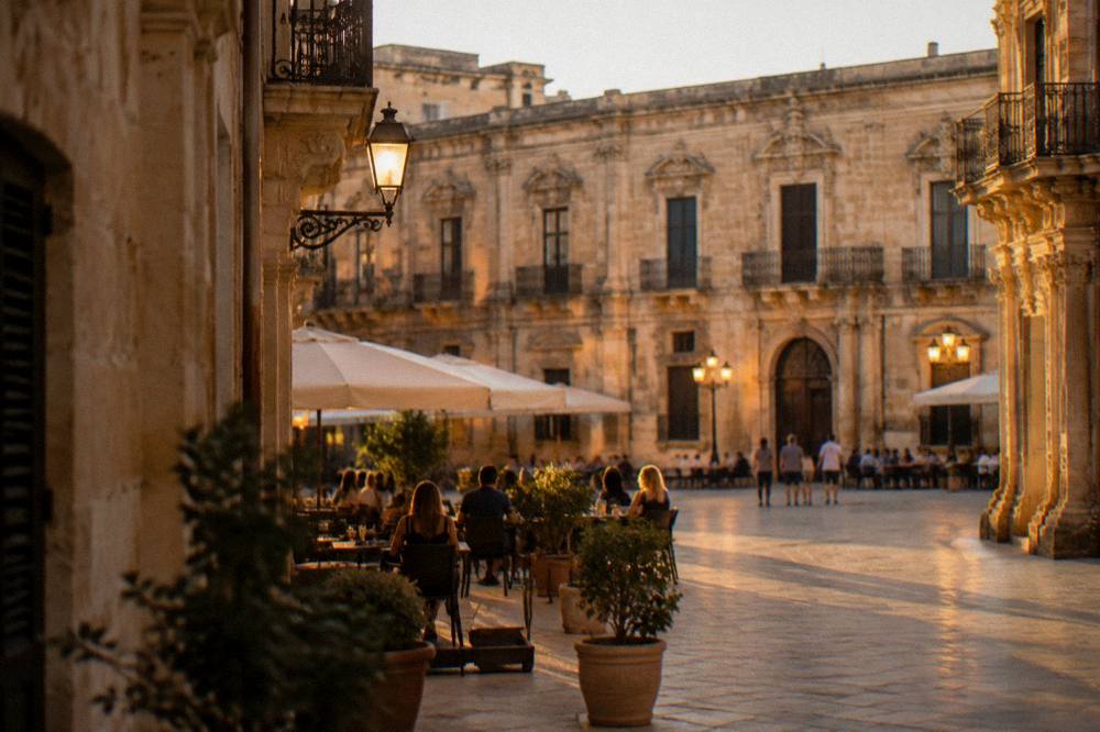 A Lecce square at dusk with warm lamplight on limestone buildings and people at outdoor tables