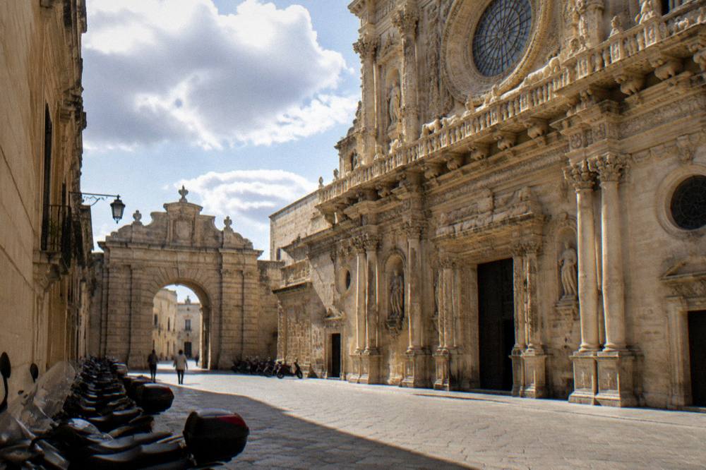 A stone city gate framed by parked scooters on a sunlit street