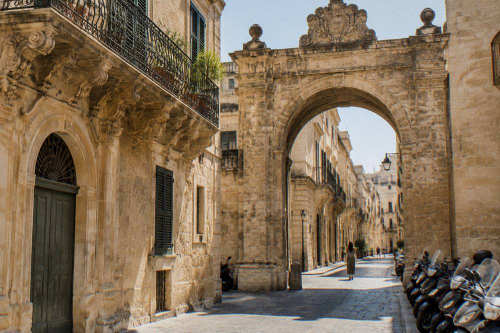 A historic stone gate in Lecce with parked scooters and a narrow street beyond