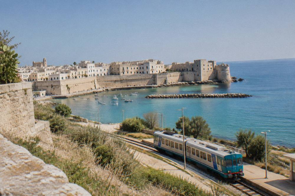 A small regional train at a rural station in Puglia under a blue sky