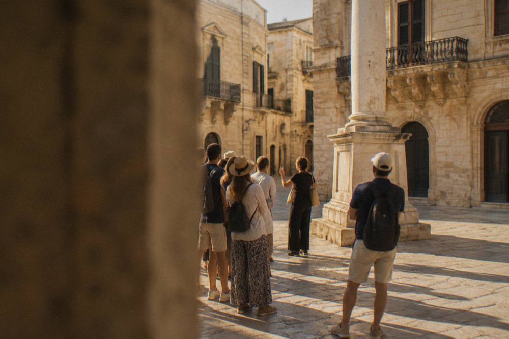 Tourists gathering around a guide in a sunlit Lecce piazza near a stone column