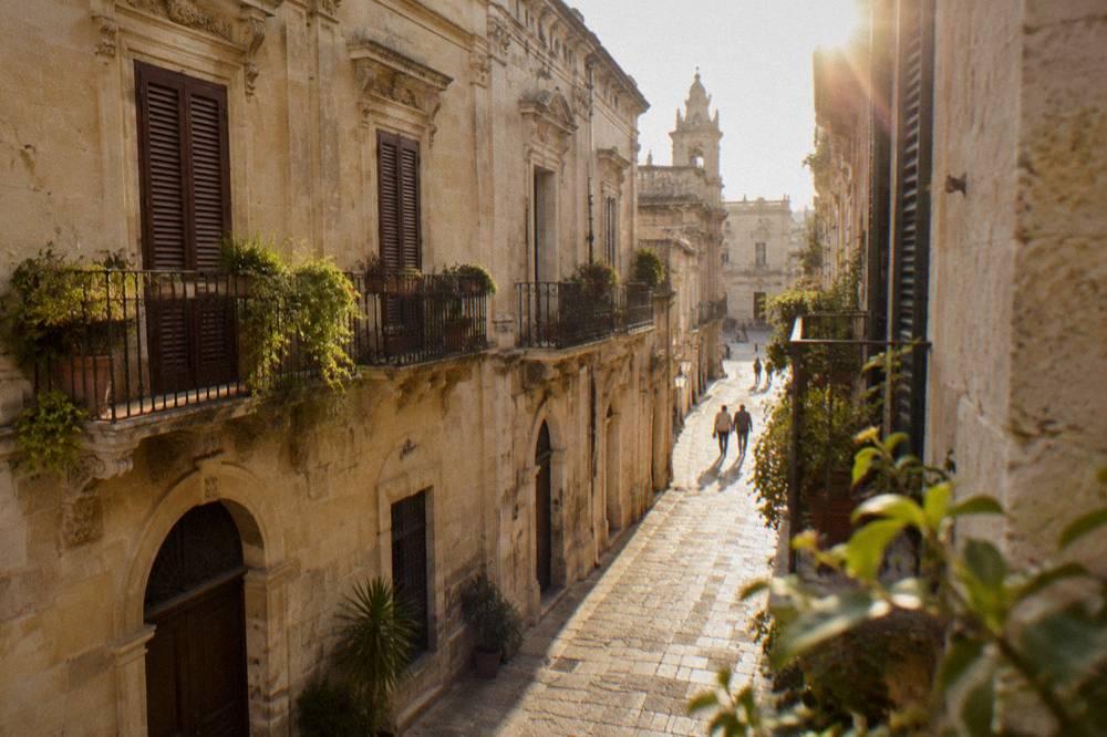 A quiet lane in Lecce with warm stone walls, plants on balconies and closed wooden shutters