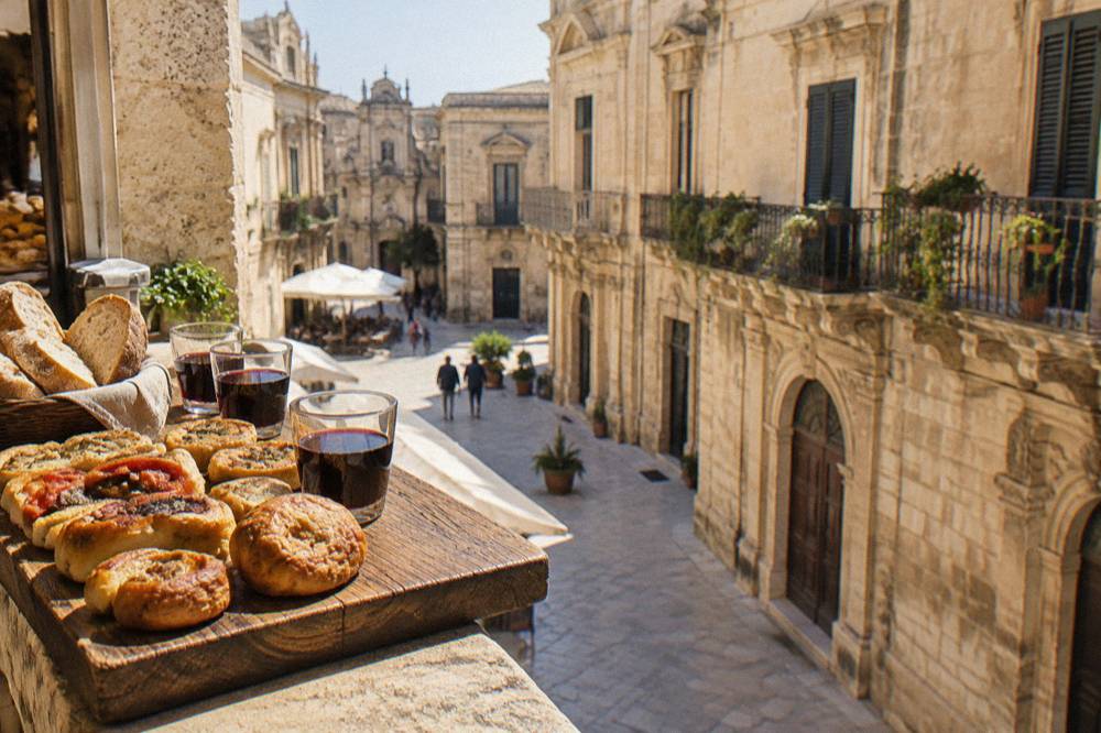 A rustic wooden board holding savoury pastries, bread and small glasses of red wine at a Lecce market counter